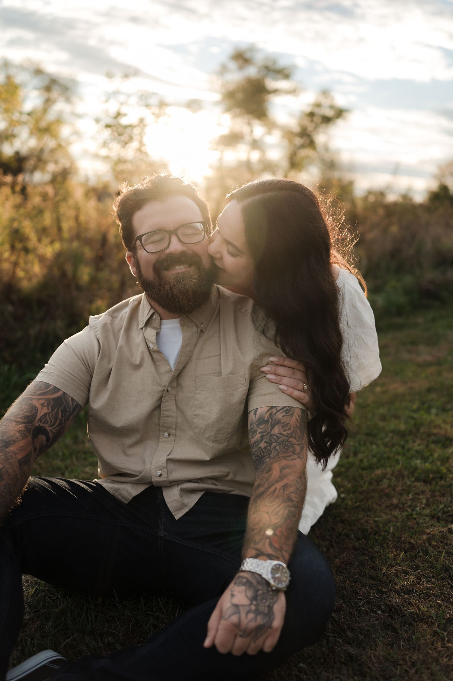 Tattooed man smiling as woman kisses his cheek while sitting on grass during a sunset engagement shoot.