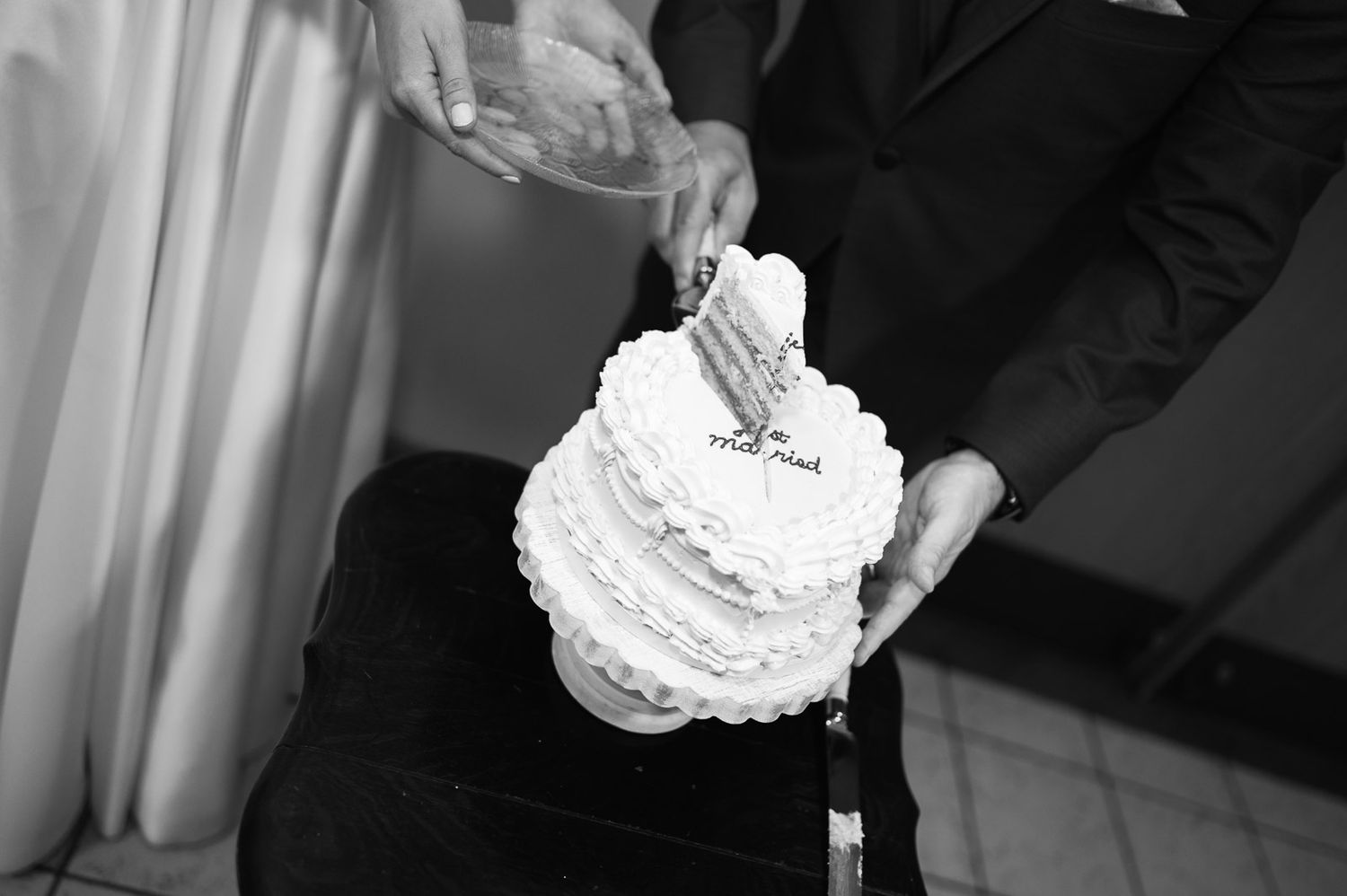 Bride and groom cutting a white tiered wedding cake with ruffled frosting in black and white photo.