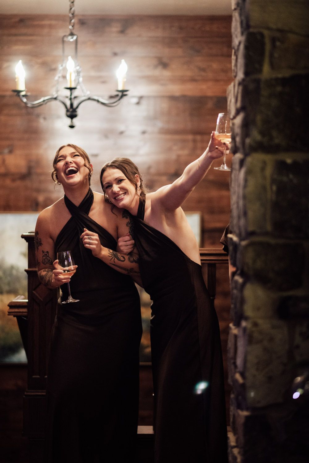 Two bridesmaids in black dresses laughing and raising champagne glasses at wedding reception.