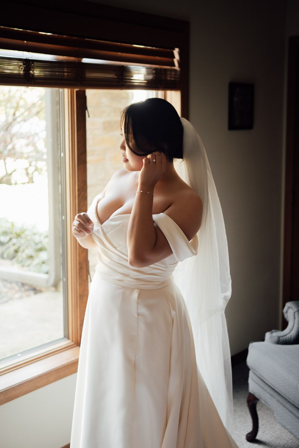 Bride in white off-shoulder wedding dress with veil looking out window while adjusting earring.