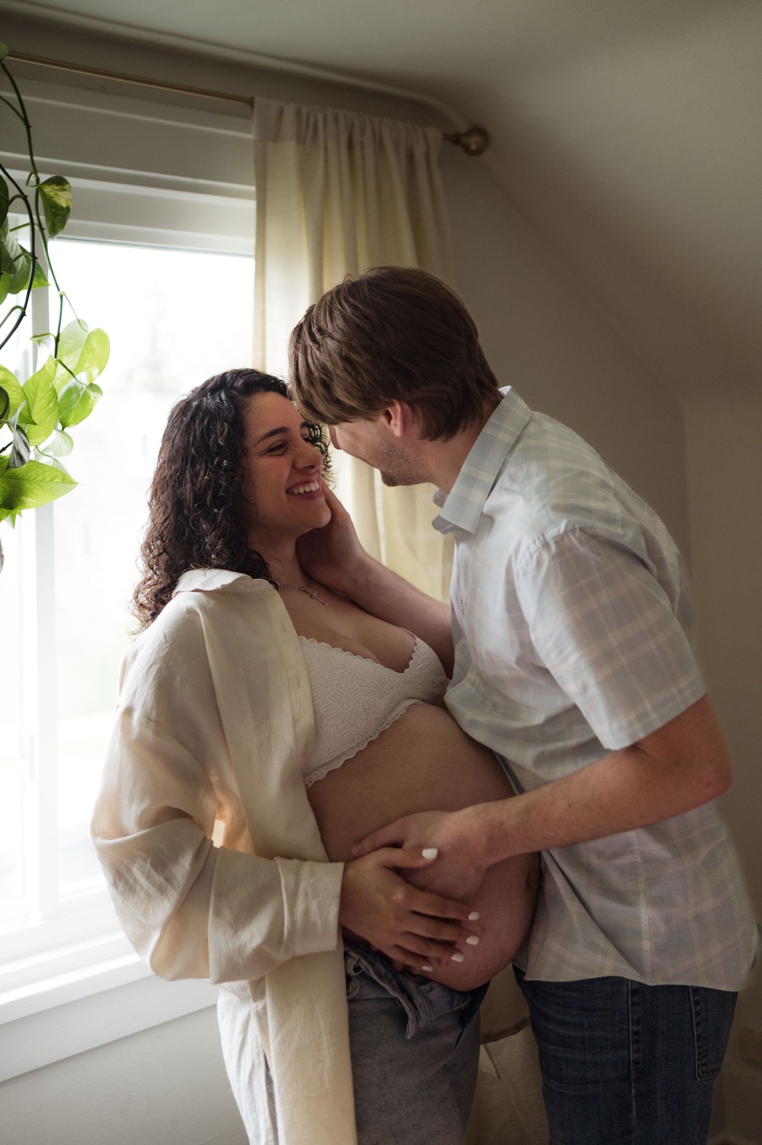 Smiling pregnant couple embracing near a bright window, man tenderly holding womans baby bump in a warmly lit nursery.