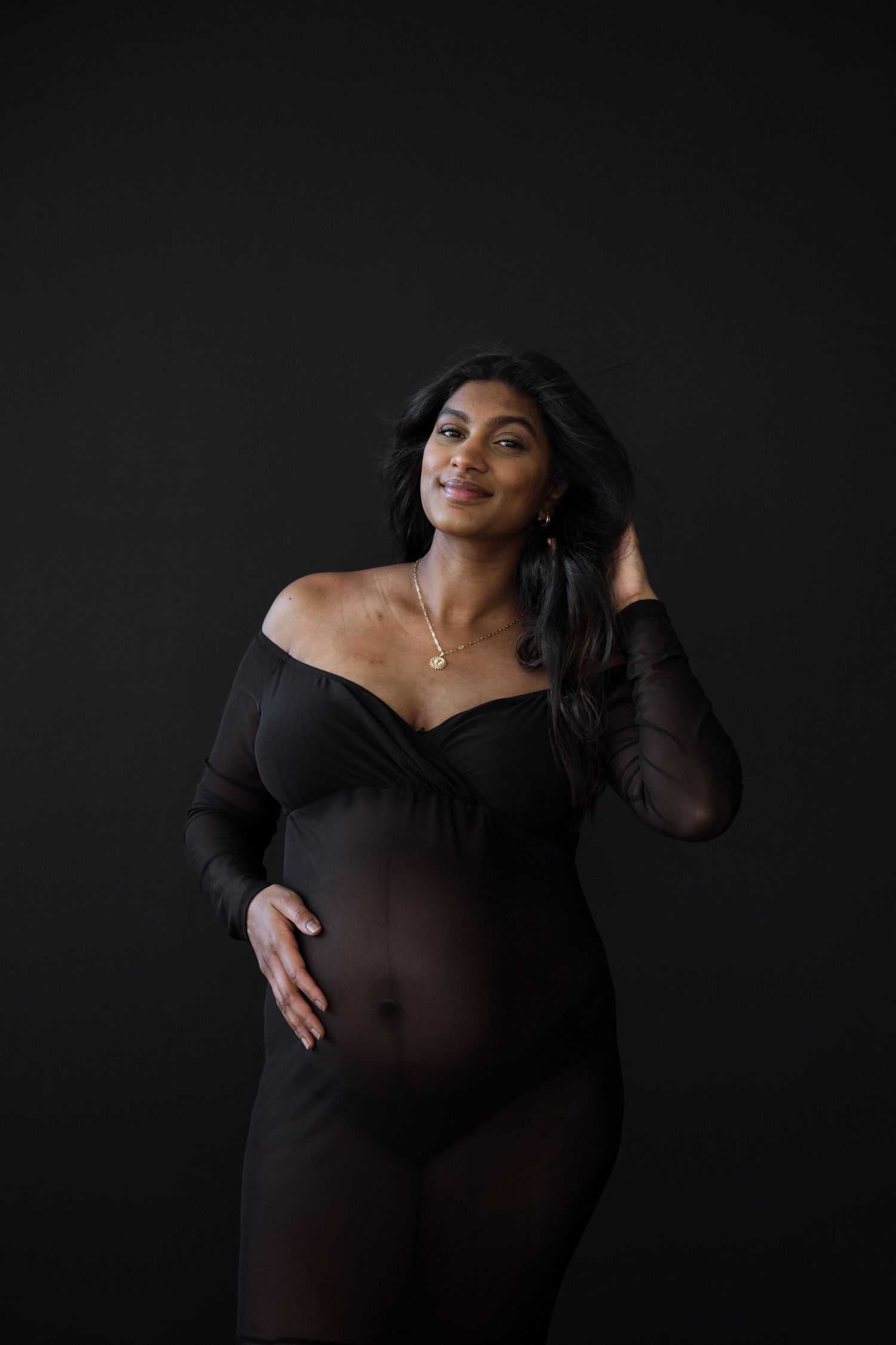 Pregnant woman in sheer black bodysuit poses against dark background smiling.