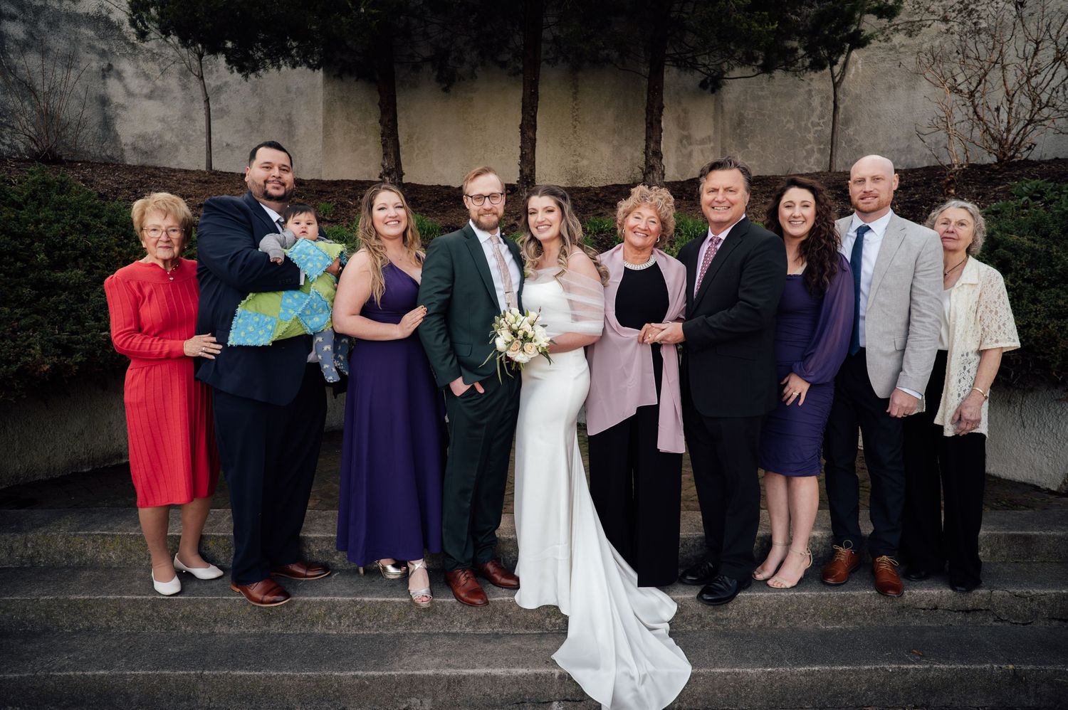 Wedding party poses outdoors on steps, bride in white gown with groom and family members in formal attire.