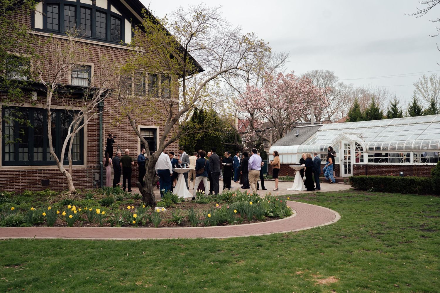Elegant outdoor wedding reception on a brick garden path beside a historic manor with a greenhouse and cherry blossom trees.