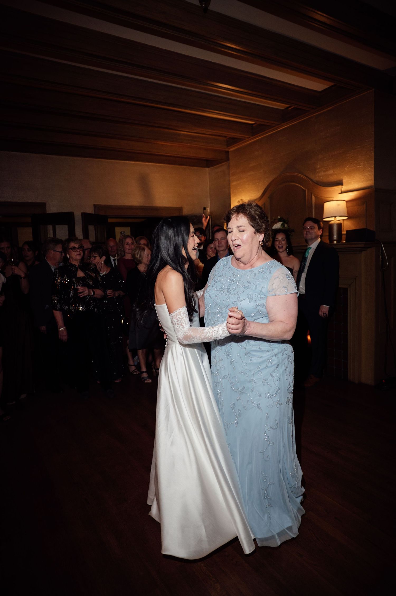 Bride in white gown dances joyfully with woman in blue lace dress at elegant wedding reception.