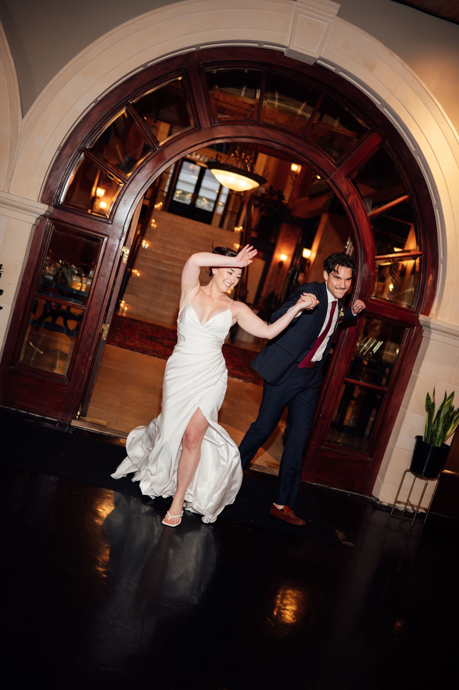 Bride and groom dancing joyfully through elegant arched wooden doors at their wedding reception.