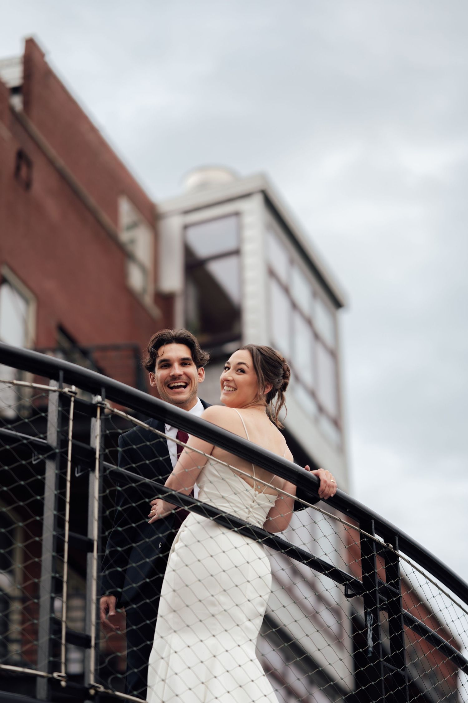 A smiling bride and groom pose on an urban rooftop staircase, with the bride in a white gown looking back joyfully.