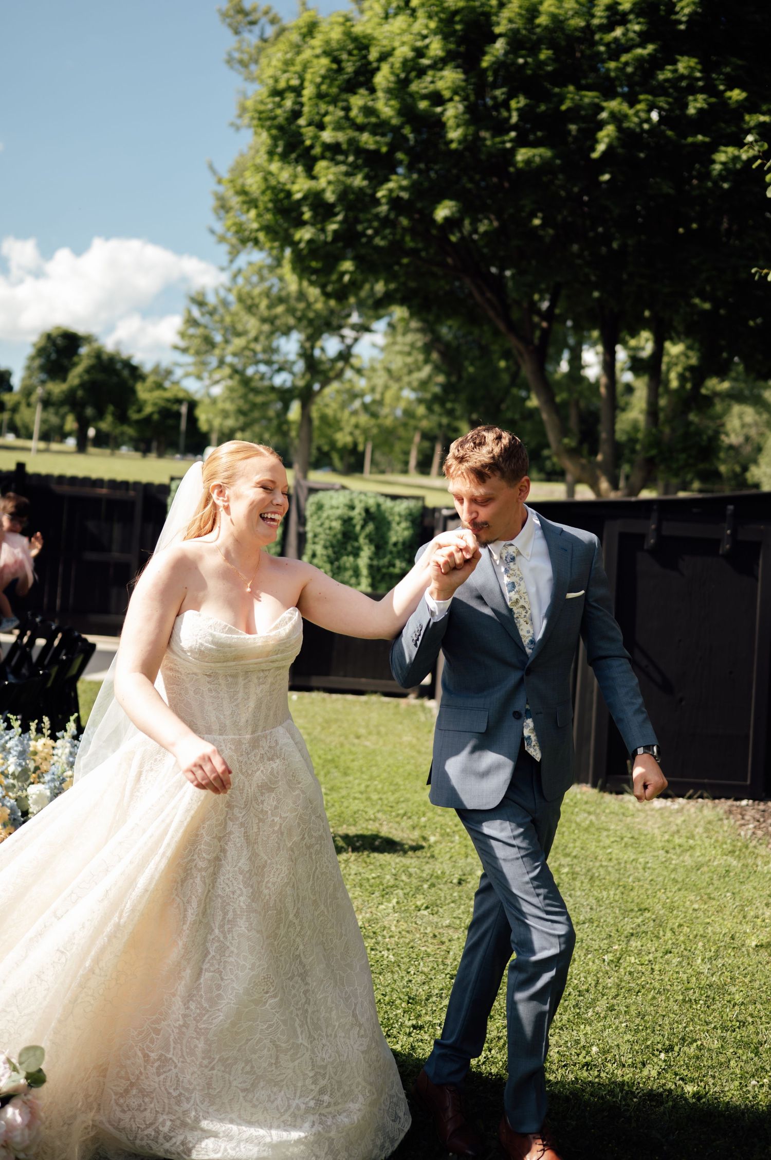 Joyful bride in lace gown and groom in navy suit walk hand in hand outdoors on sunny wedding day.