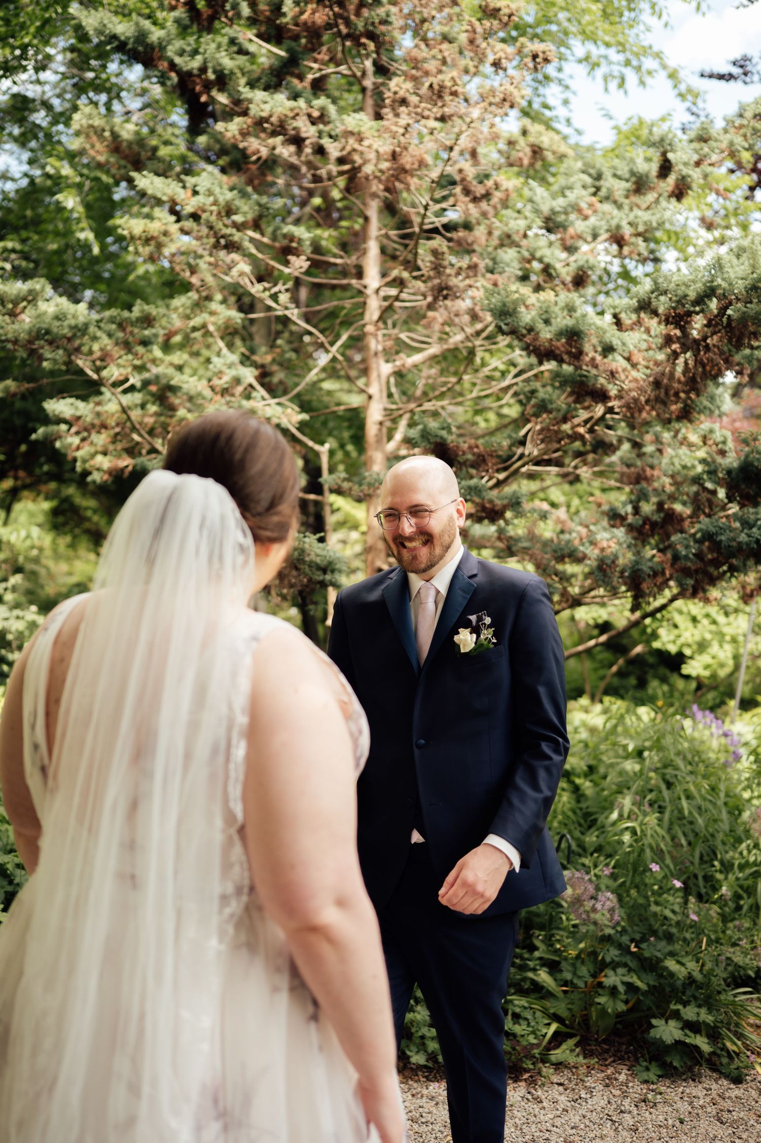 Groom in navy suit smiles at bride in white wedding gown and veil during outdoor first look in lush garden.