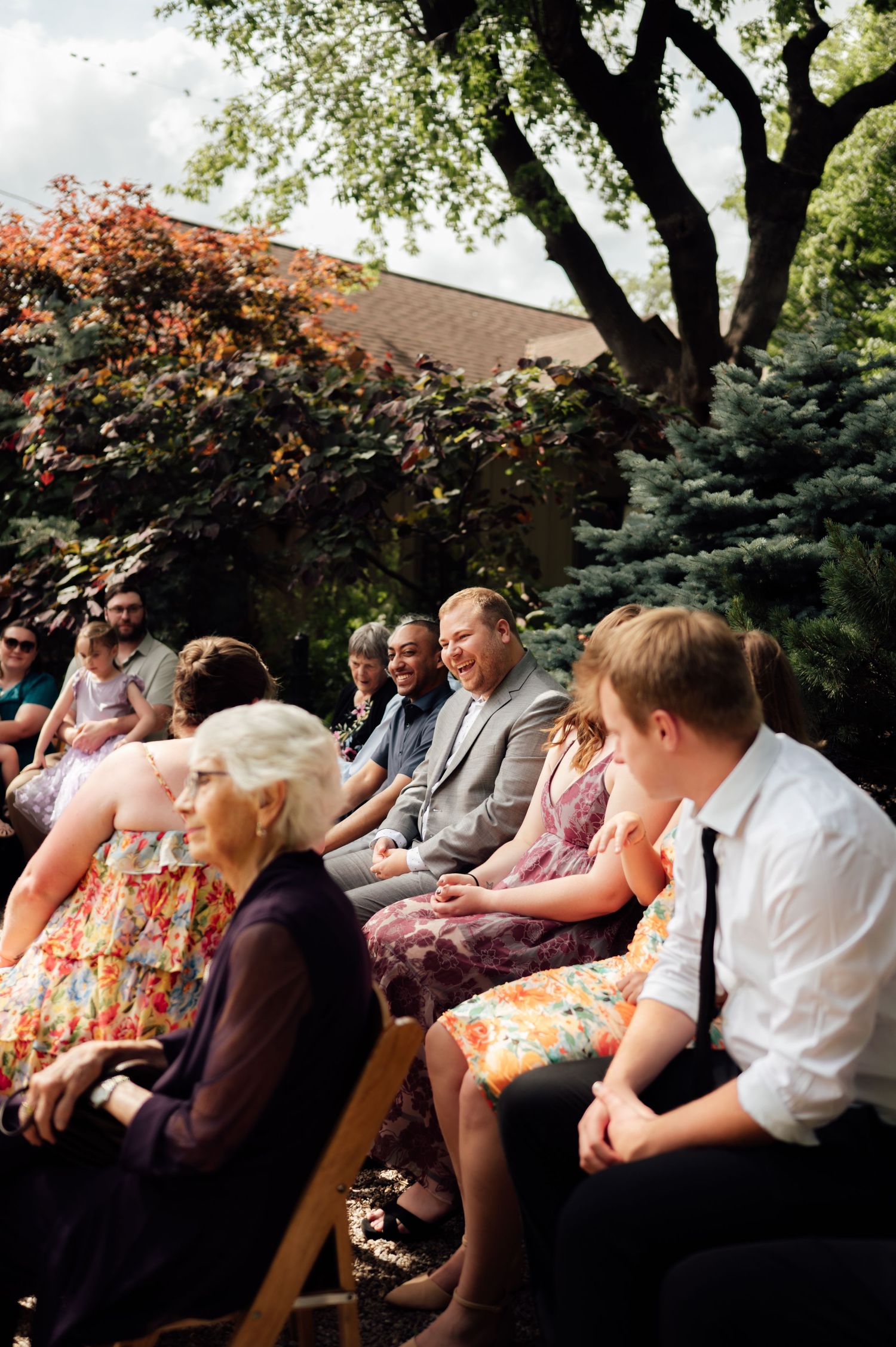 Guests laugh and chat at an outdoor wedding ceremony, seated on wooden chairs surrounded by lush green trees.