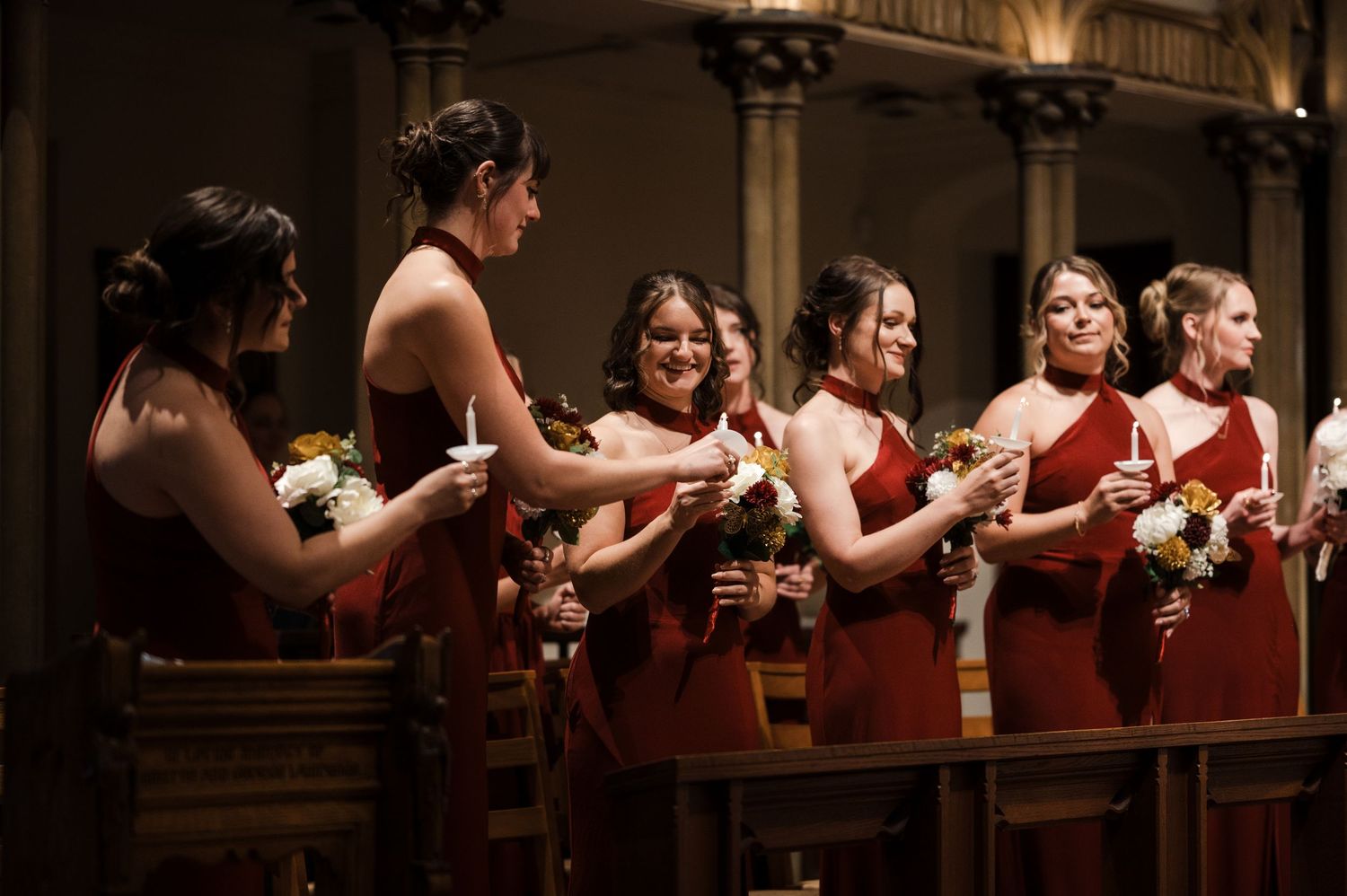 Bridesmaids in burgundy dresses stand in a row holding bouquets at an elegant indoor wedding venue.