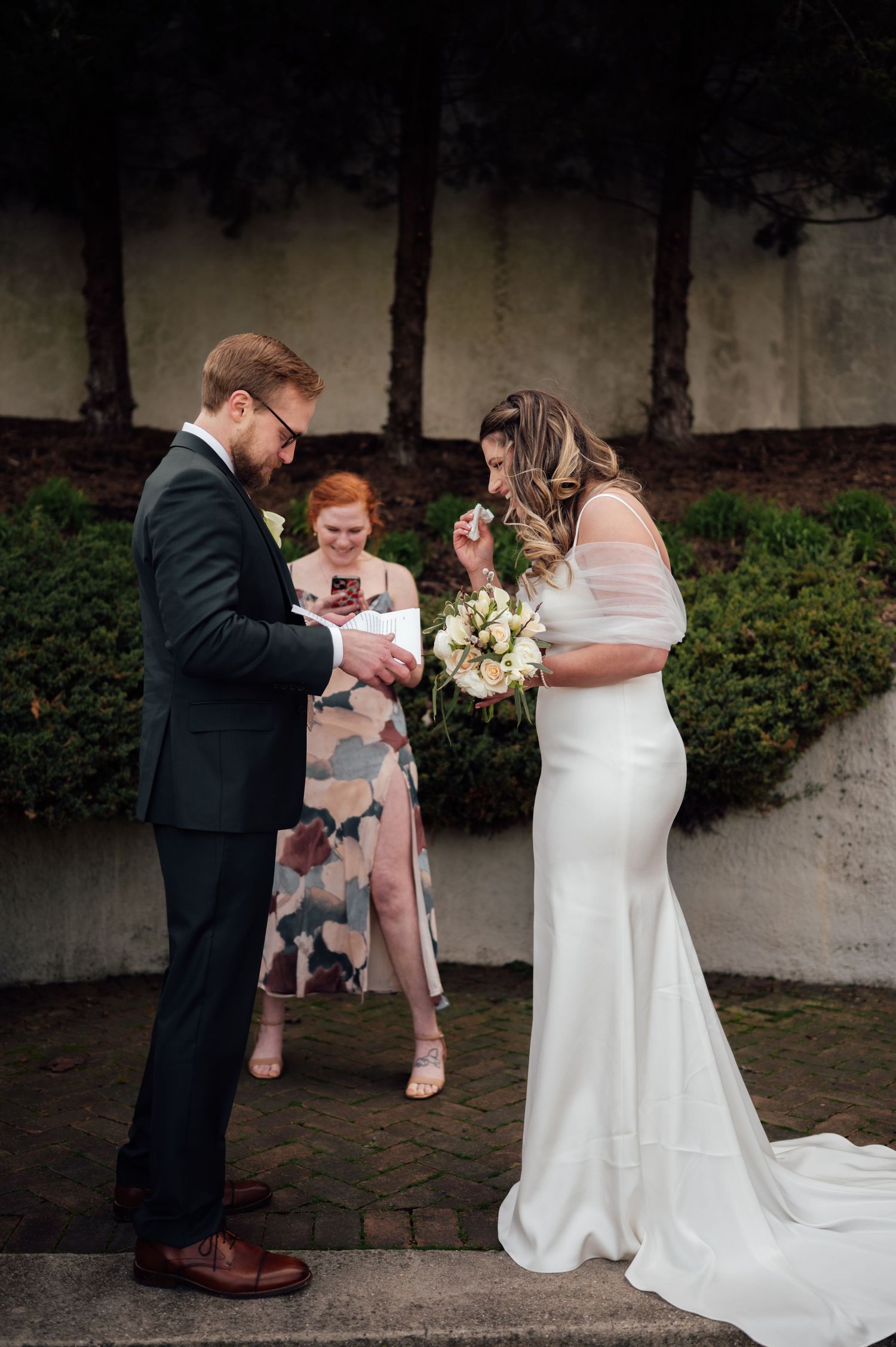 Bride and groom exchange rings during outdoor wedding ceremony, officiant stands behind them.