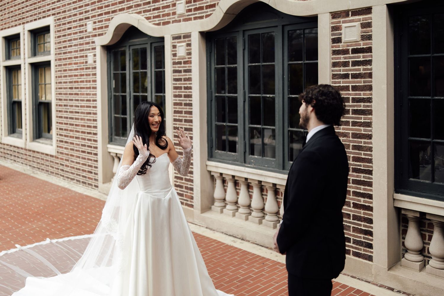 Bride in white gown with long veil shares emotional first look moment with groom in black suit outside brick building.