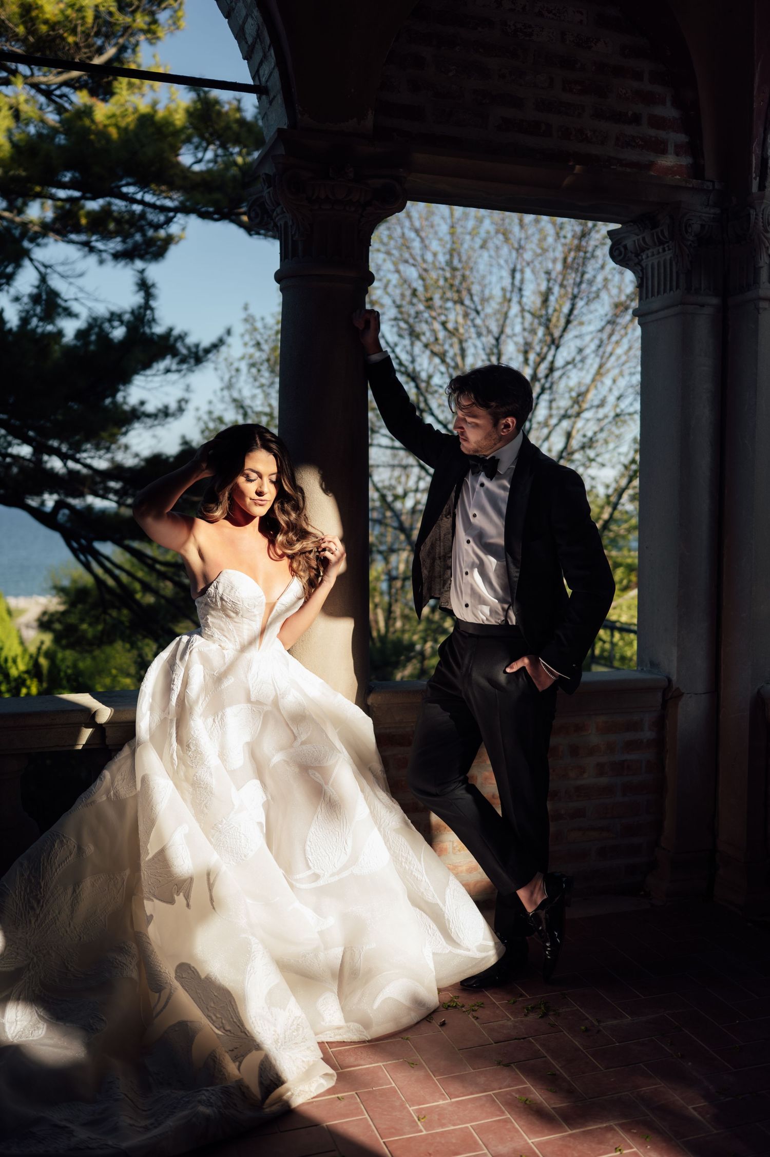 Bride in ballgown and groom in tuxedo posing dramatically on sunlit brick terrace with scenic backdrop.