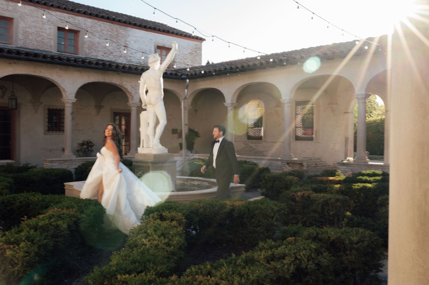 Bride and groom walking past classical statue fountain in elegant courtyard with string lights at sunset.