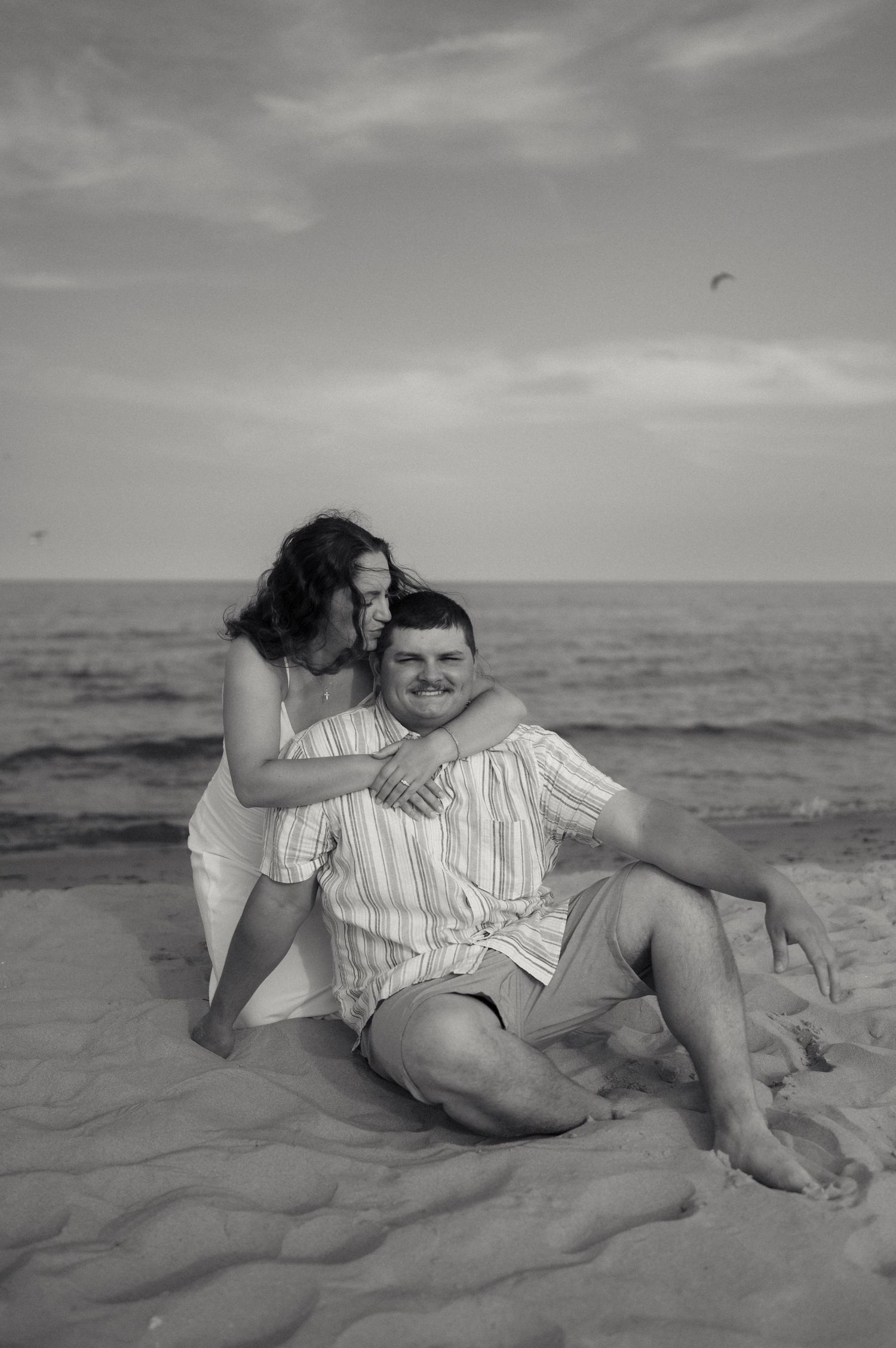 Black and white photo of couple sitting on beach sand with woman embracing man from behind.