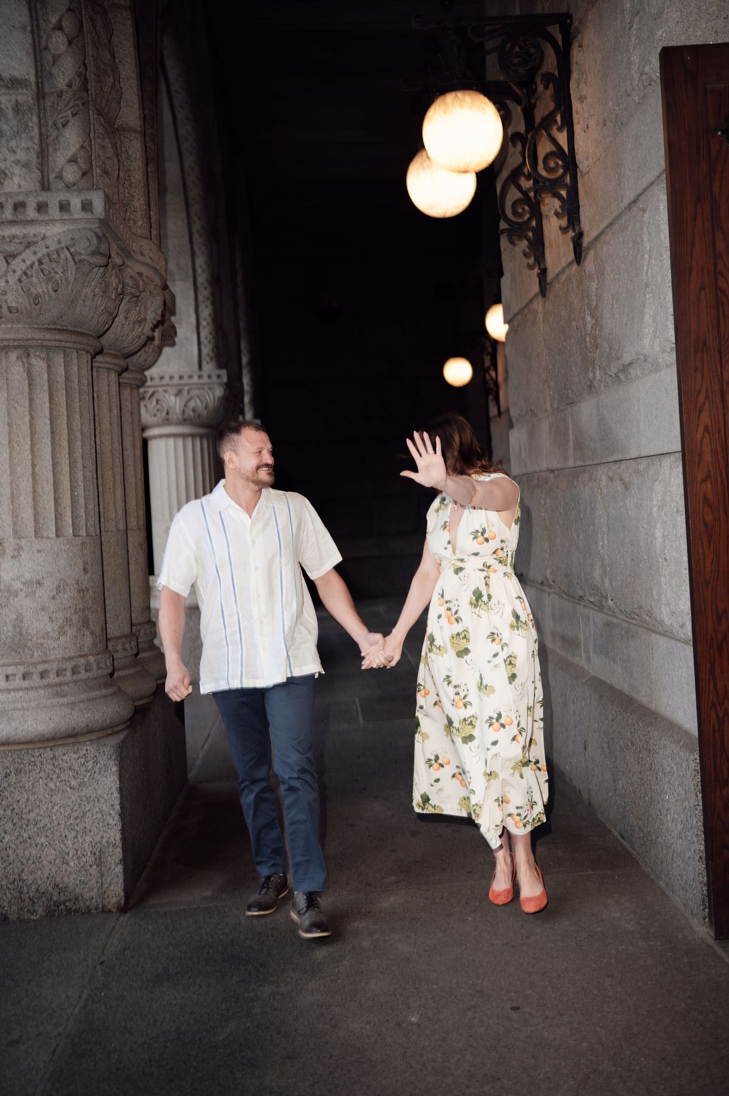 Couple holding hands walking through a grand stone archway with glowing globe lights overhead.