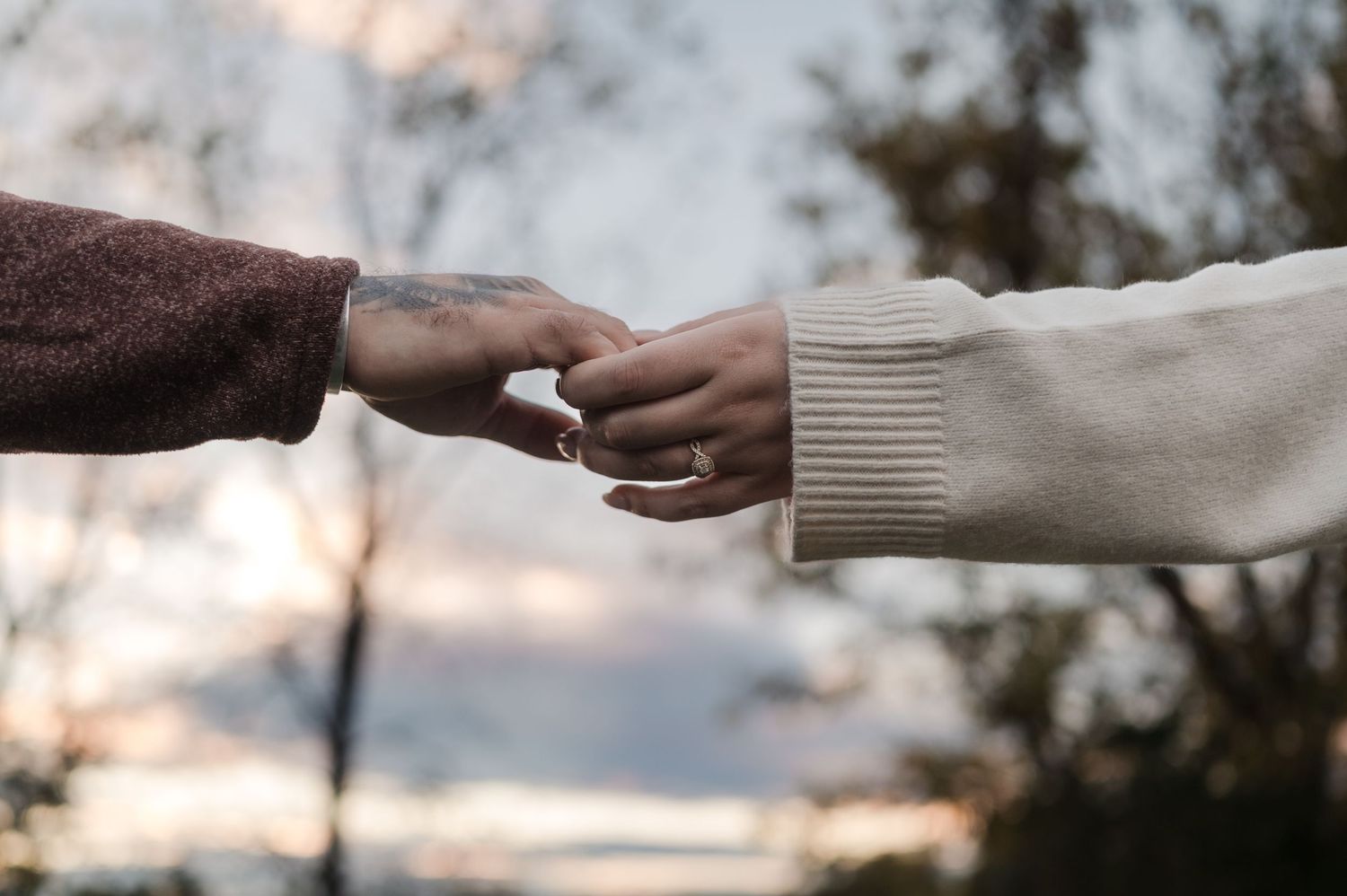 Close-up of couple holding hands at sunset, woman wearing engagement ring on white sweater sleeve.