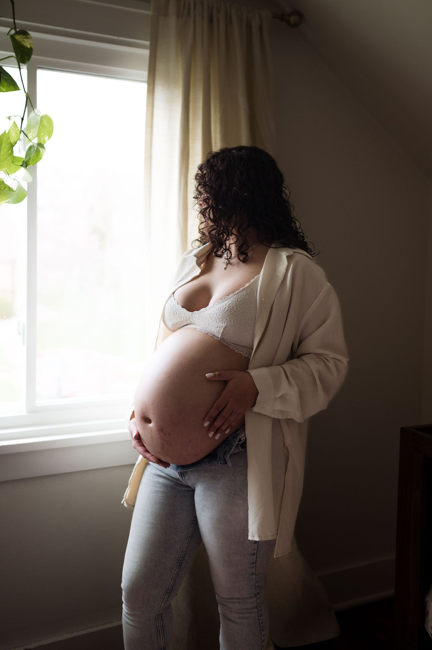 Pregnant woman in white cardigan and lace bralette gazing out a window while cradling her bare baby bump in soft light.