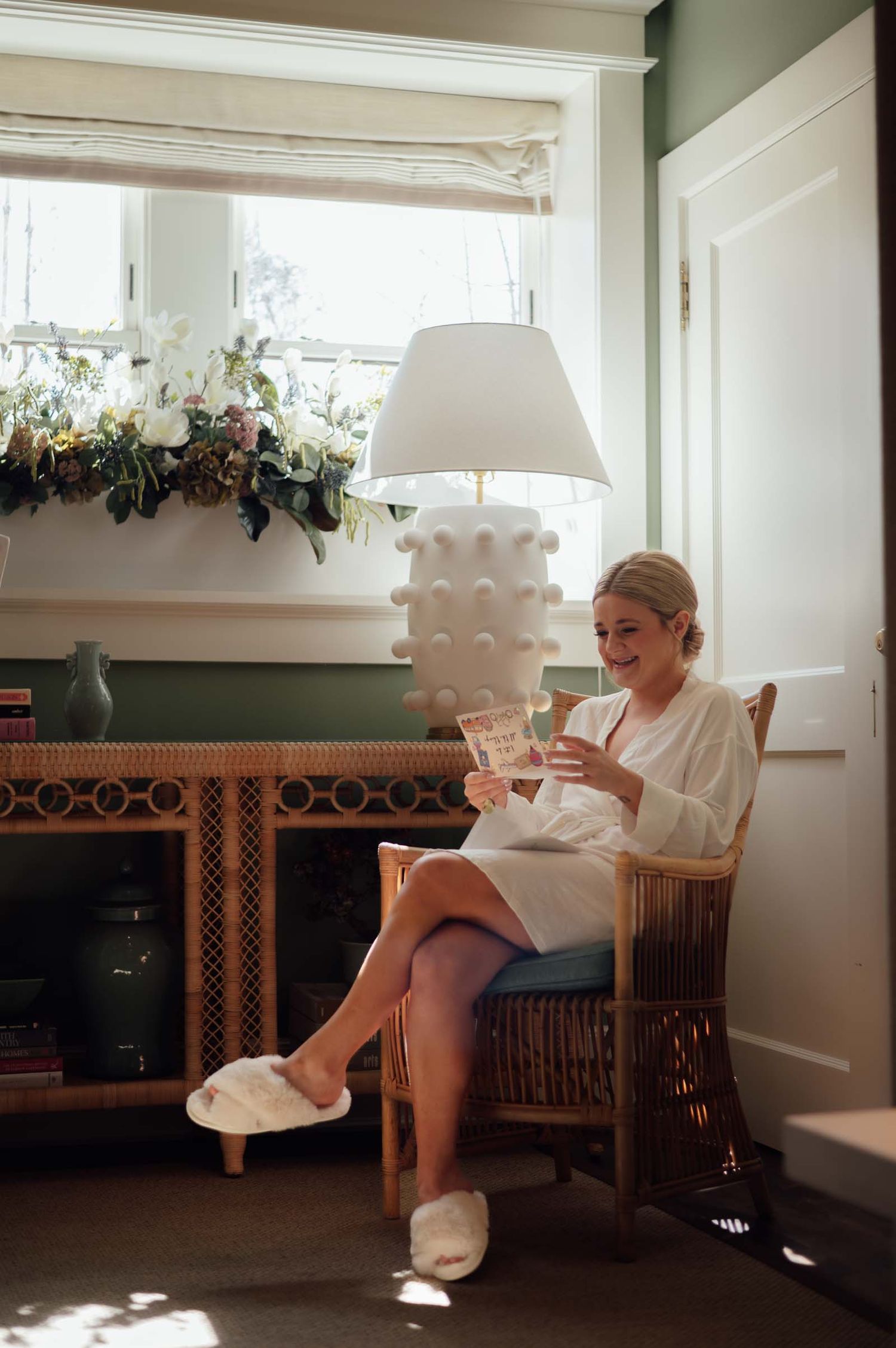 Blonde bride in white robe reading card while sitting in wicker chair by lamp-lit window with flowers.