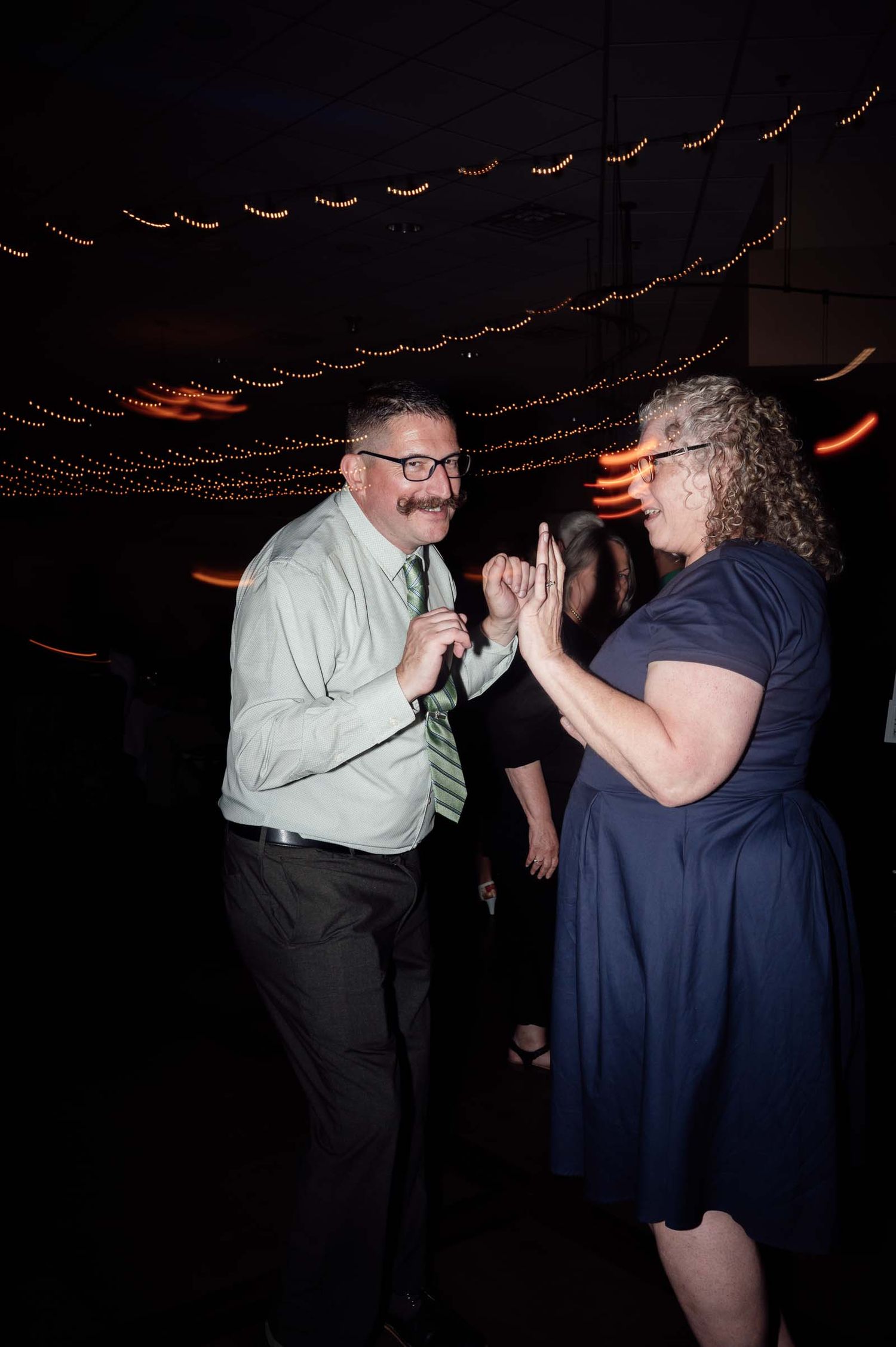 Joyful wedding guests in formal attire dancing and laughing together under string lights at evening reception.