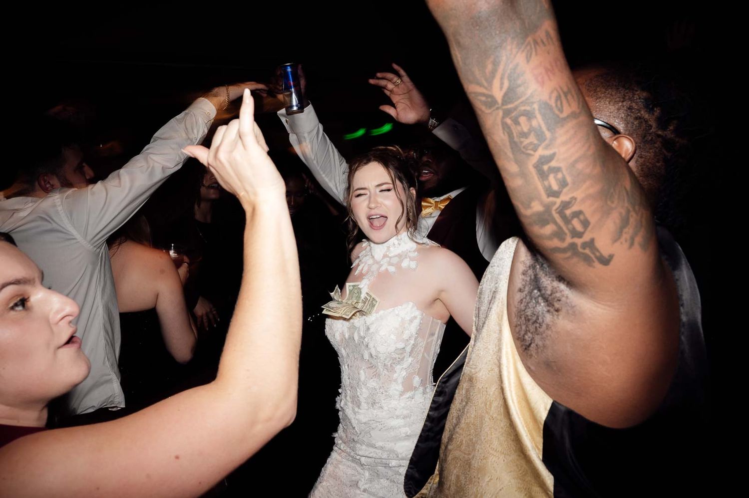 Bride in white lace gown dances joyfully with guests on a dark wedding reception dance floor.