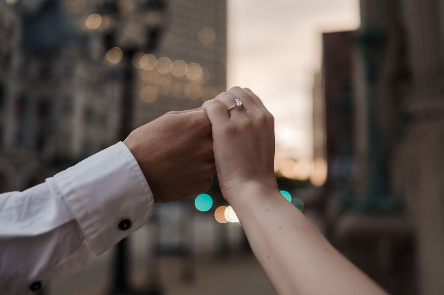 Close-up of couple holding hands with engagement ring visible against city bokeh background.