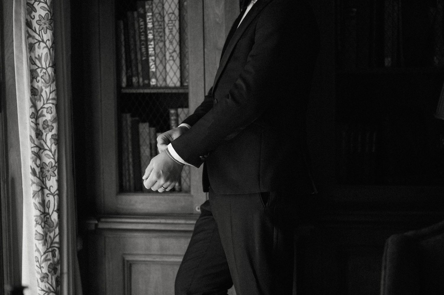 A man in a black suit stands near a bookshelf in a dimly lit room, captured in black and white.