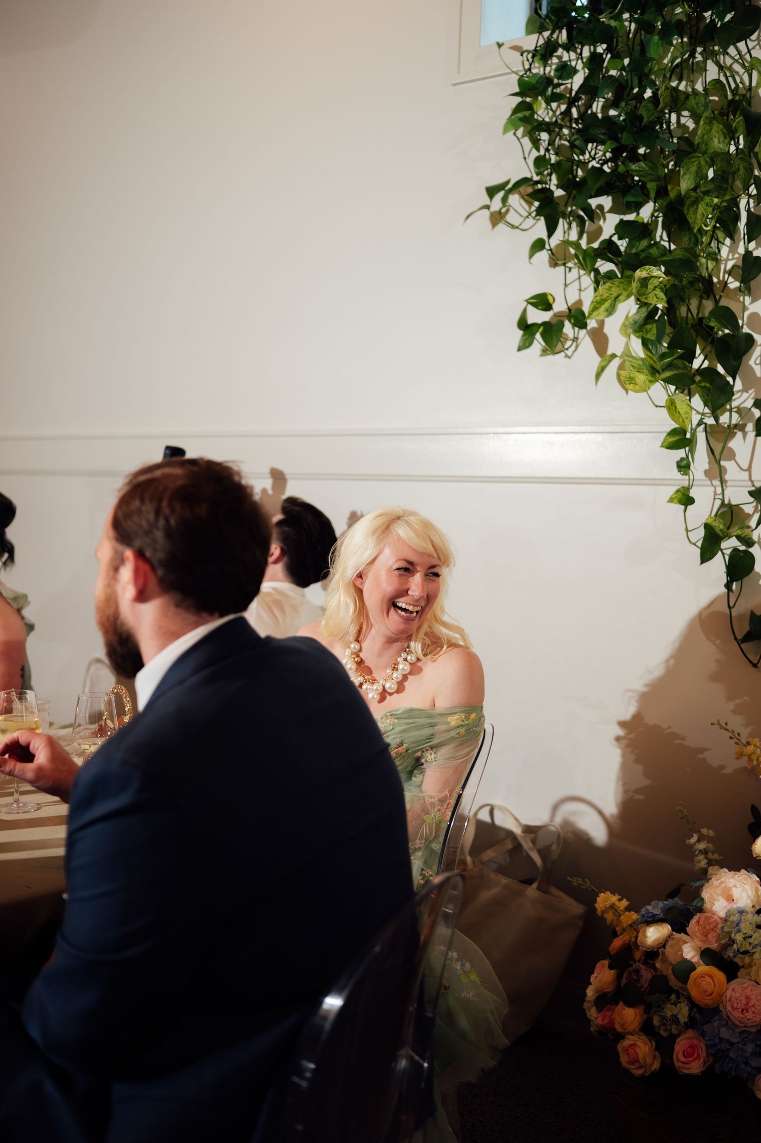 Blonde woman in green off-shoulder dress laughing at elegant wedding reception table with floral arrangements.
