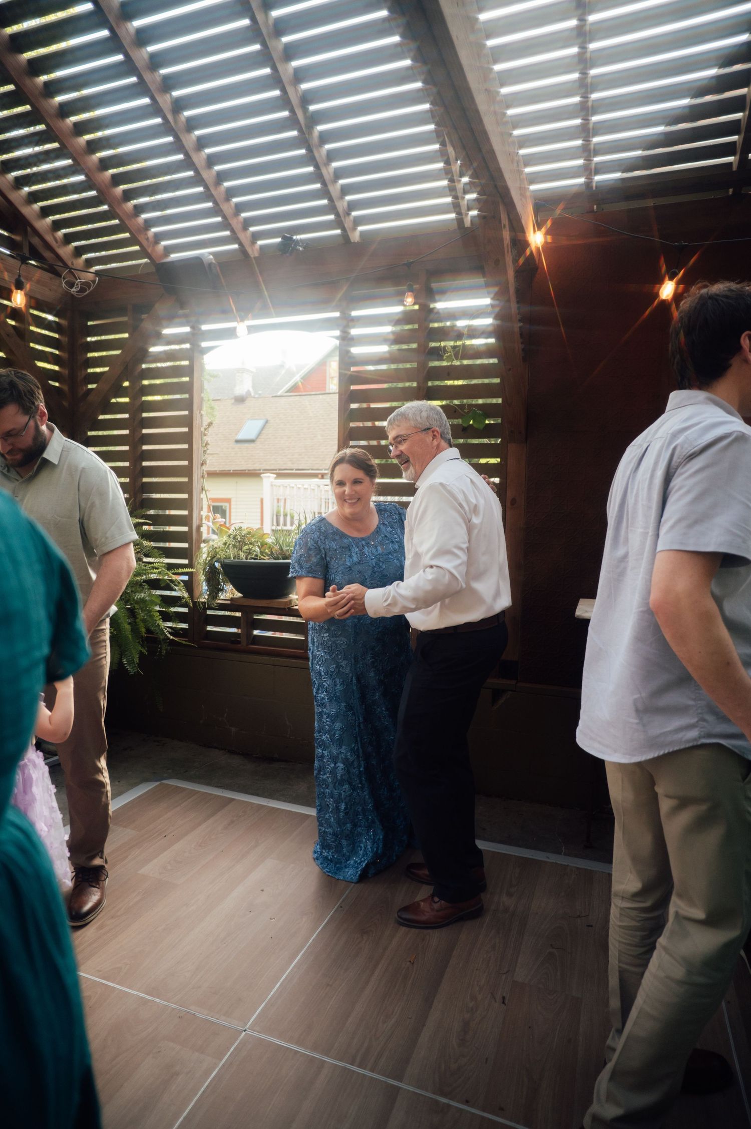 Older couple dancing together at an evening outdoor reception under a pergola with warm string lights.