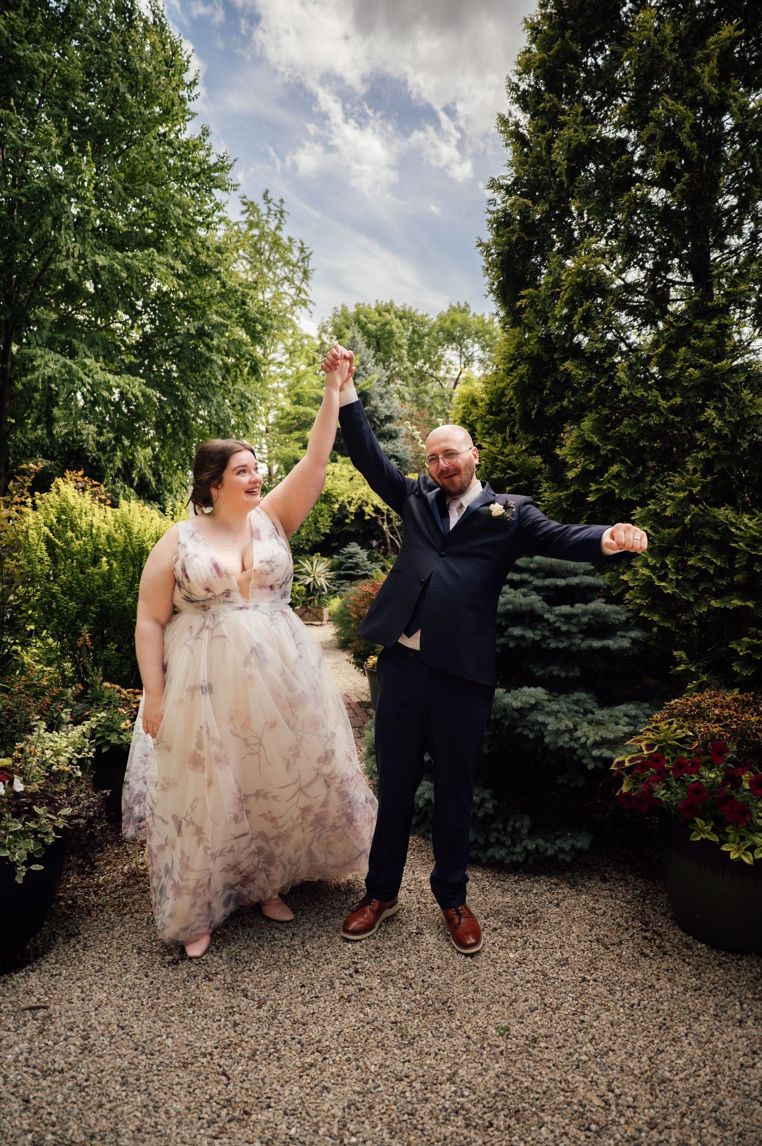 Joyful bride and groom celebrate in a lush garden, holding hands raised high in their wedding attire.