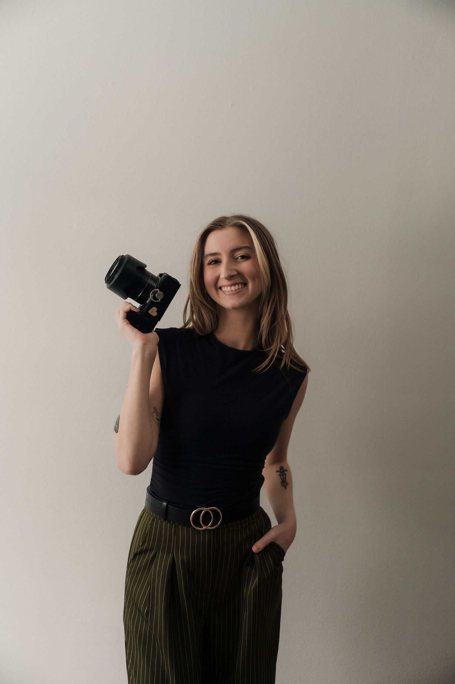 Smiling female photographer holding DSLR camera up in black top and olive striped trousers.