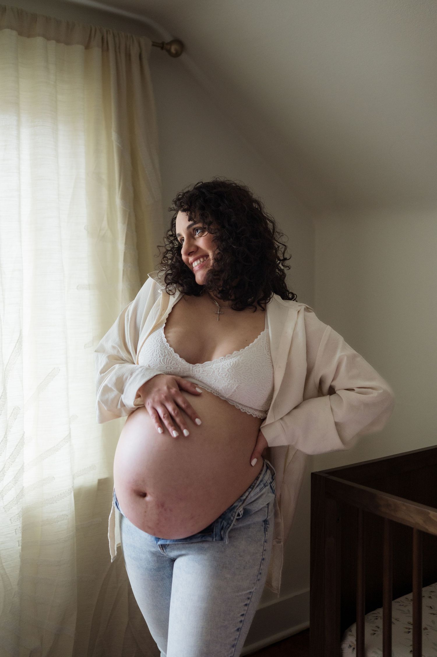 A pregnant woman in jeans and white cardigan stands by a window in natural light, gently touching her baby bump.