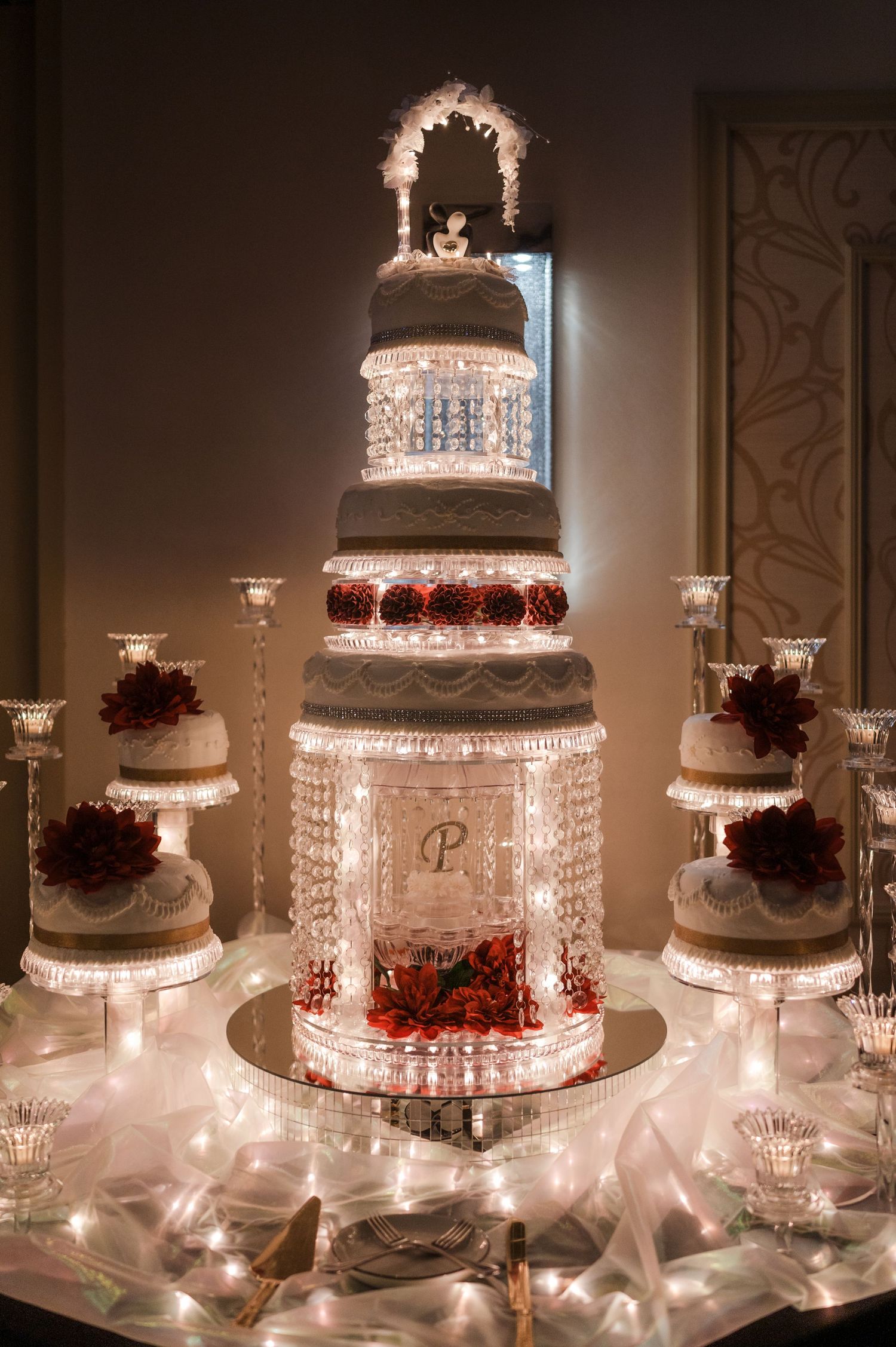 Elegant multi-tiered wedding cake with crystal stands and red floral accents illuminated by soft lighting.