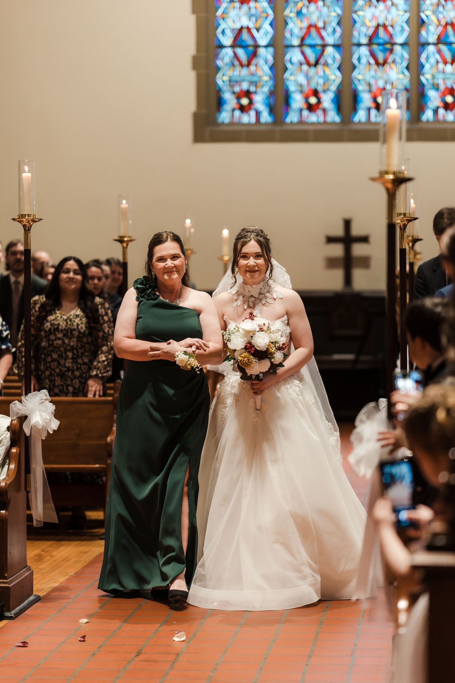 Wedding party members walk down church aisle during ceremony with candles and stained glass windows in background.
