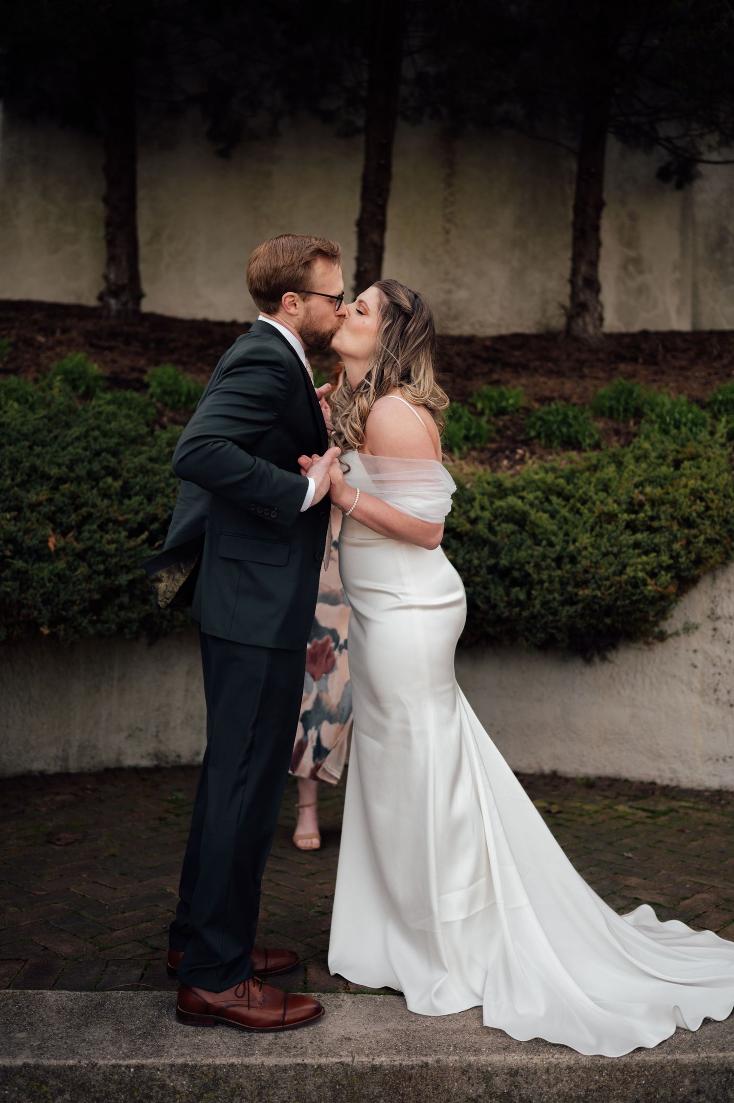 A couple shares a romantic kiss outdoors, bride in white off-shoulder gown, groom in dark suit, on their wedding day.