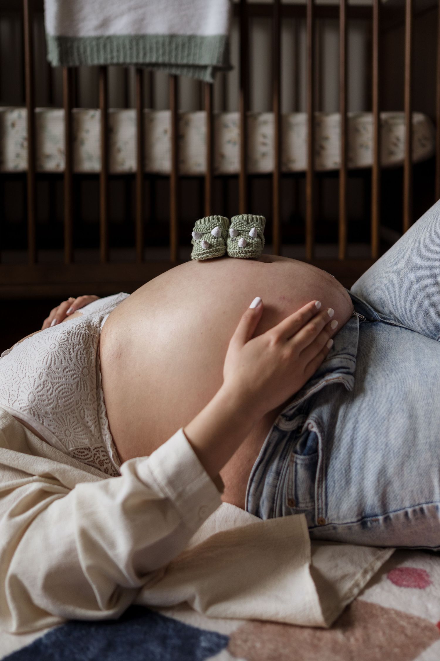 Close-up of pregnant belly with tiny baby booties balanced on top, lying near a wooden crib in soft natural light.