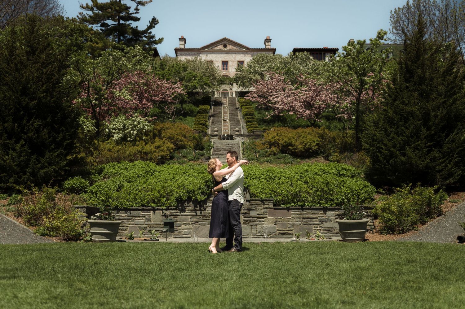 Couple embracing on manicured lawn before a grand estate with blooming cherry trees and stone steps.