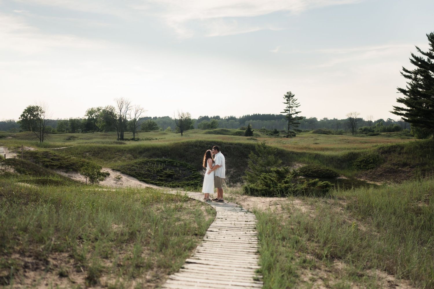 Couple standing together on a wooden boardwalk path through scenic sandy dunes and green grass.