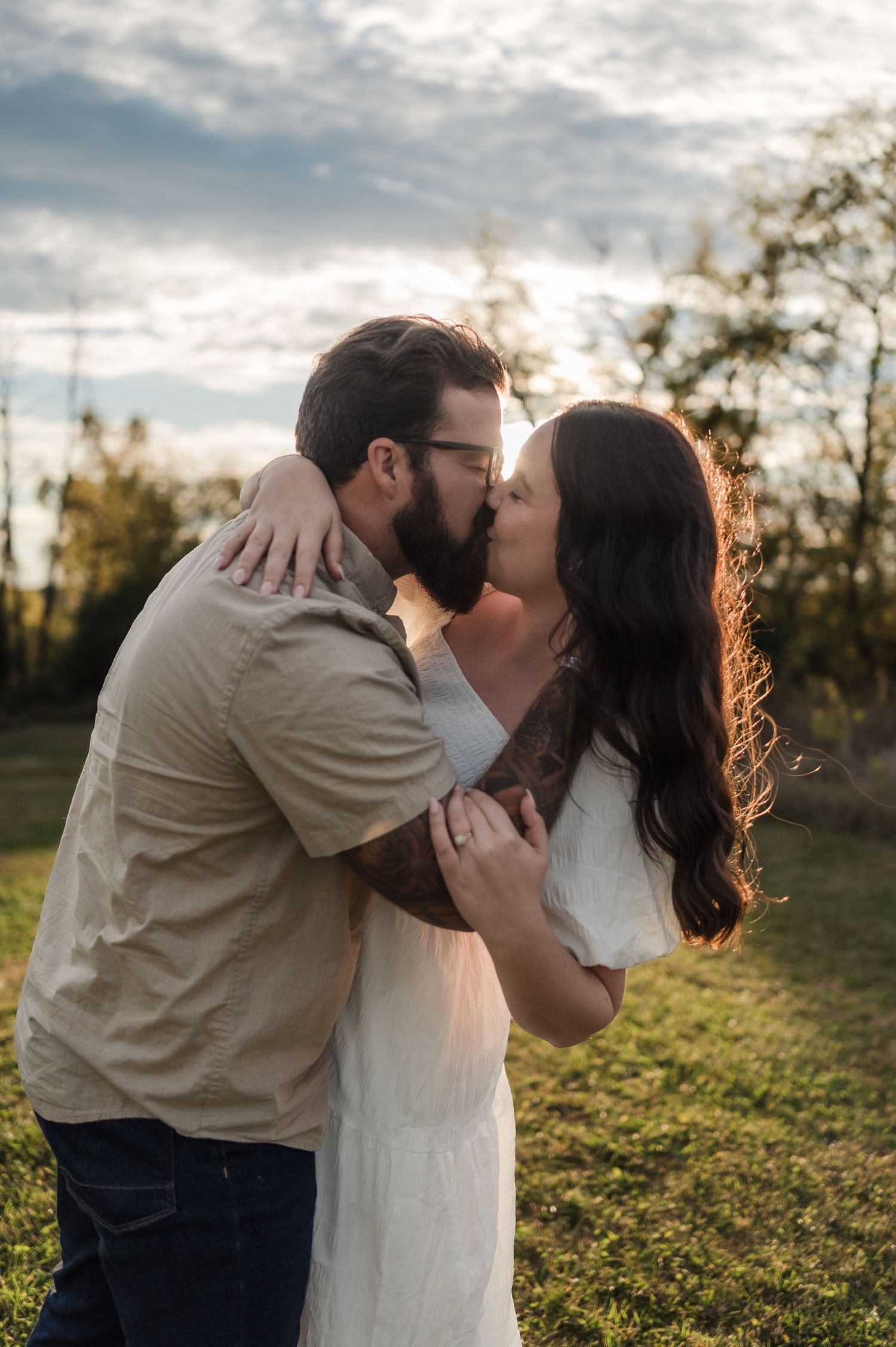 Tattooed bearded man kissing brunette woman in a white dress during golden hour outdoor engagement session.