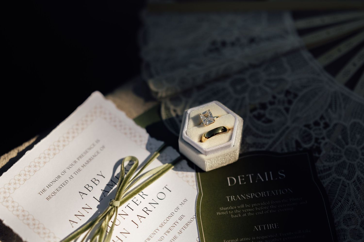 Wedding details including gold rings in a velvet box, invitations, and dark stationery on a dark surface.