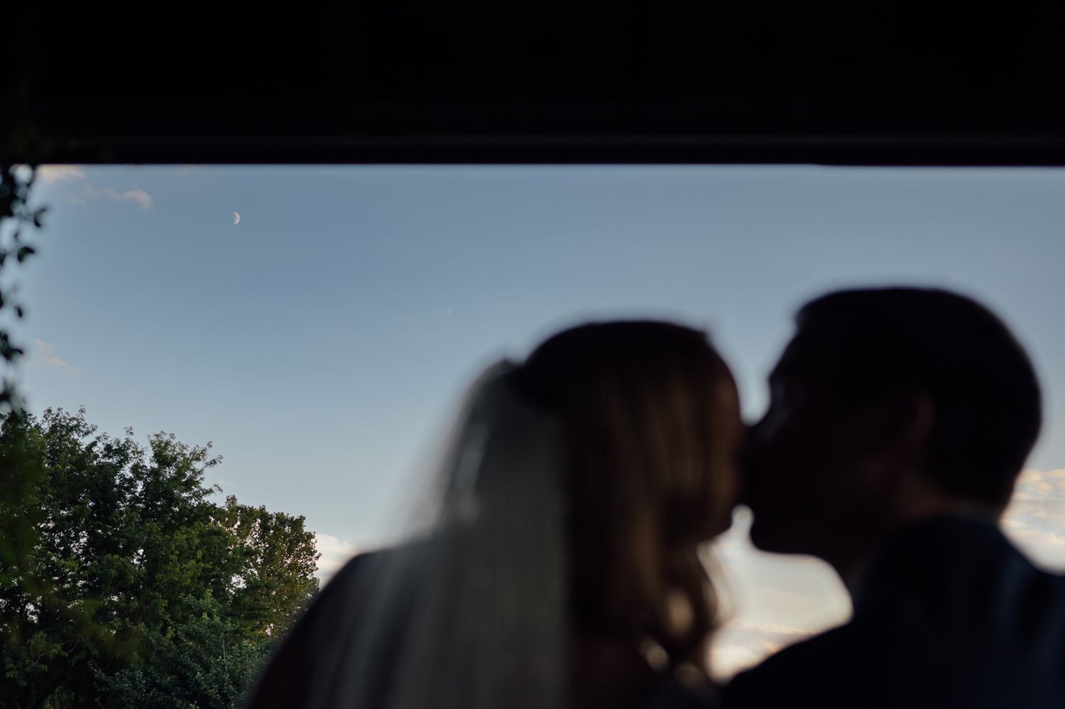 Silhouette of bride and groom sharing a romantic kiss at dusk framed by a window.