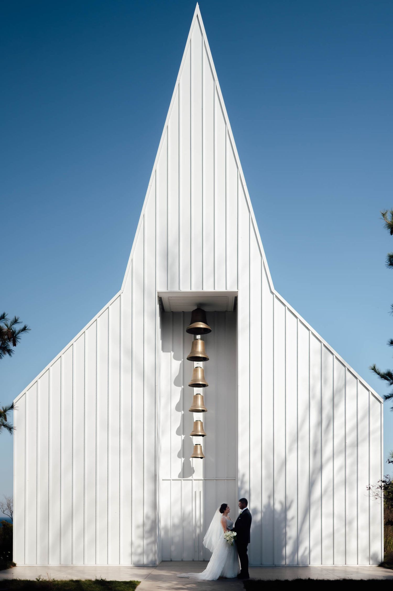 Bride and groom pose romantically before striking modern white triangular chapel with decorative bells outside.