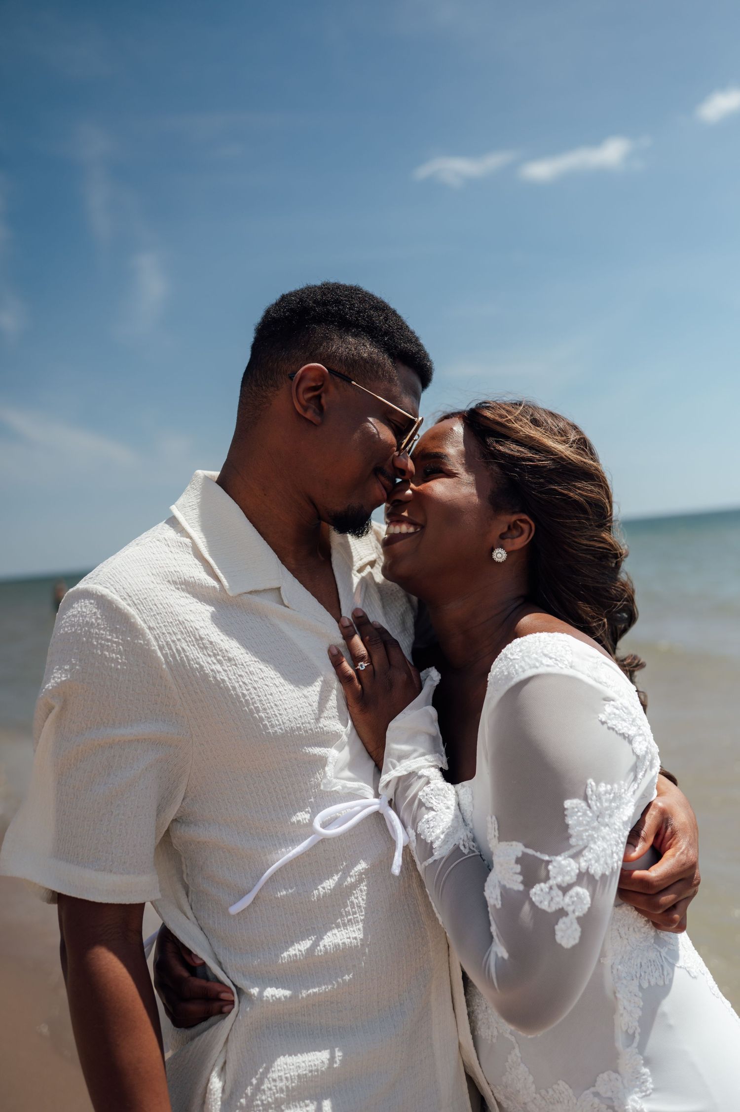 A joyful couple in white attire shares a romantic moment on a sunny beach, nearly kissing with the ocean behind them.