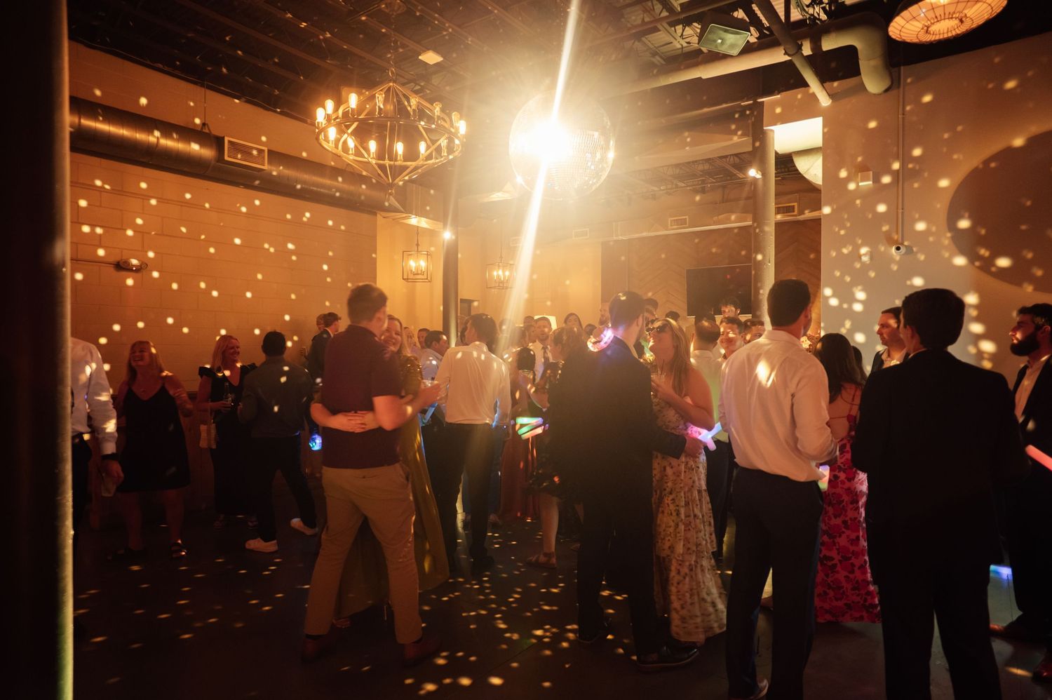 Guests dancing at a dimly lit wedding reception with glowing lights and chandelier ambiance.