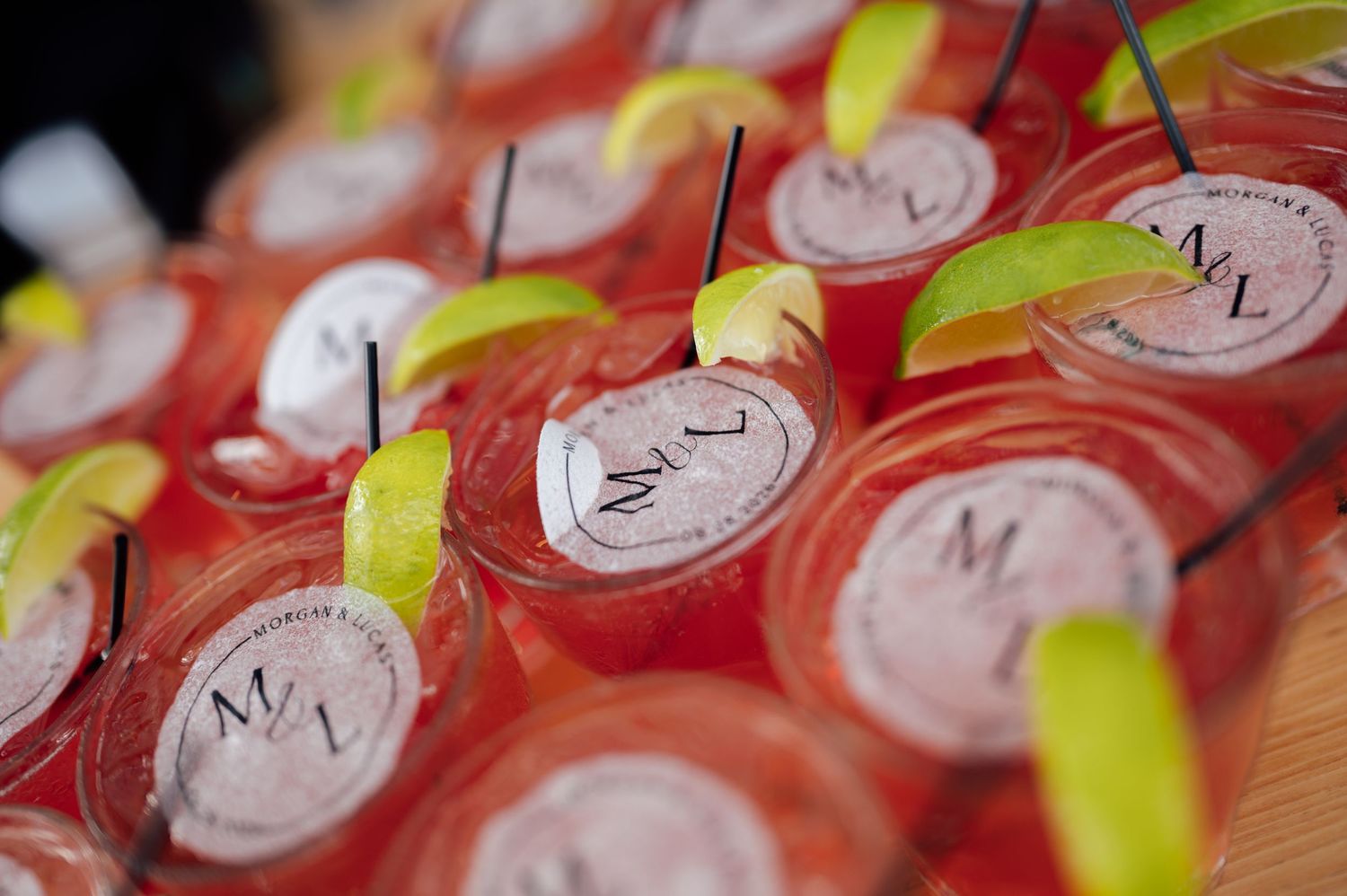 Rows of pink cocktails in branded cups garnished with lime wedges and black straws at a catered event.