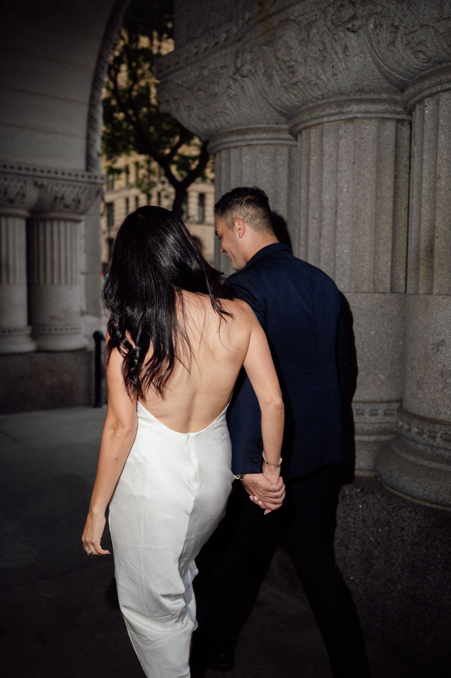 Couple holding hands walking past ornate stone columns, woman in backless white dress and man in navy suit.