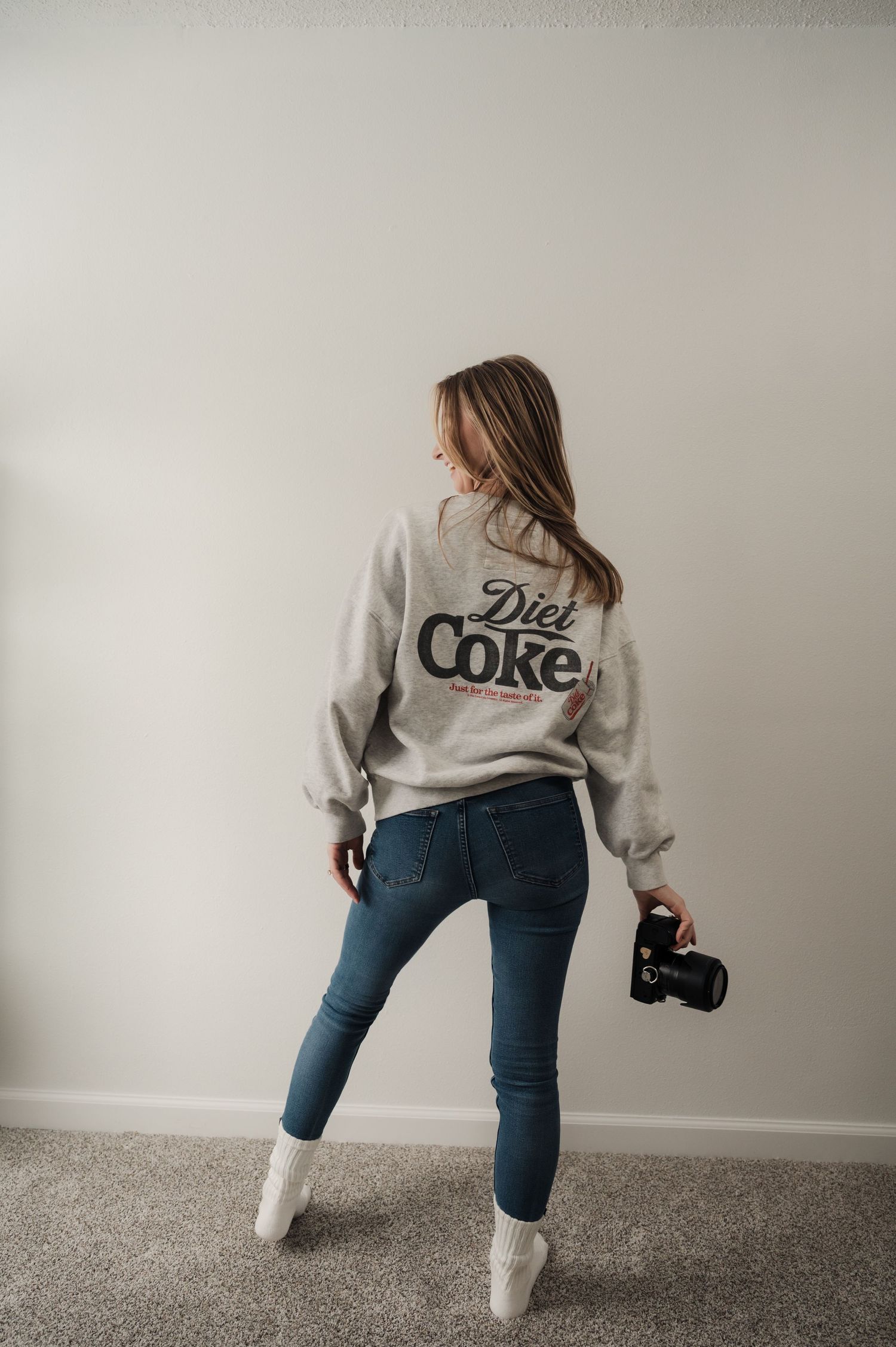 Woman in Diet Coke sweatshirt and jeans holding a camera, posed with back to camera against white wall.