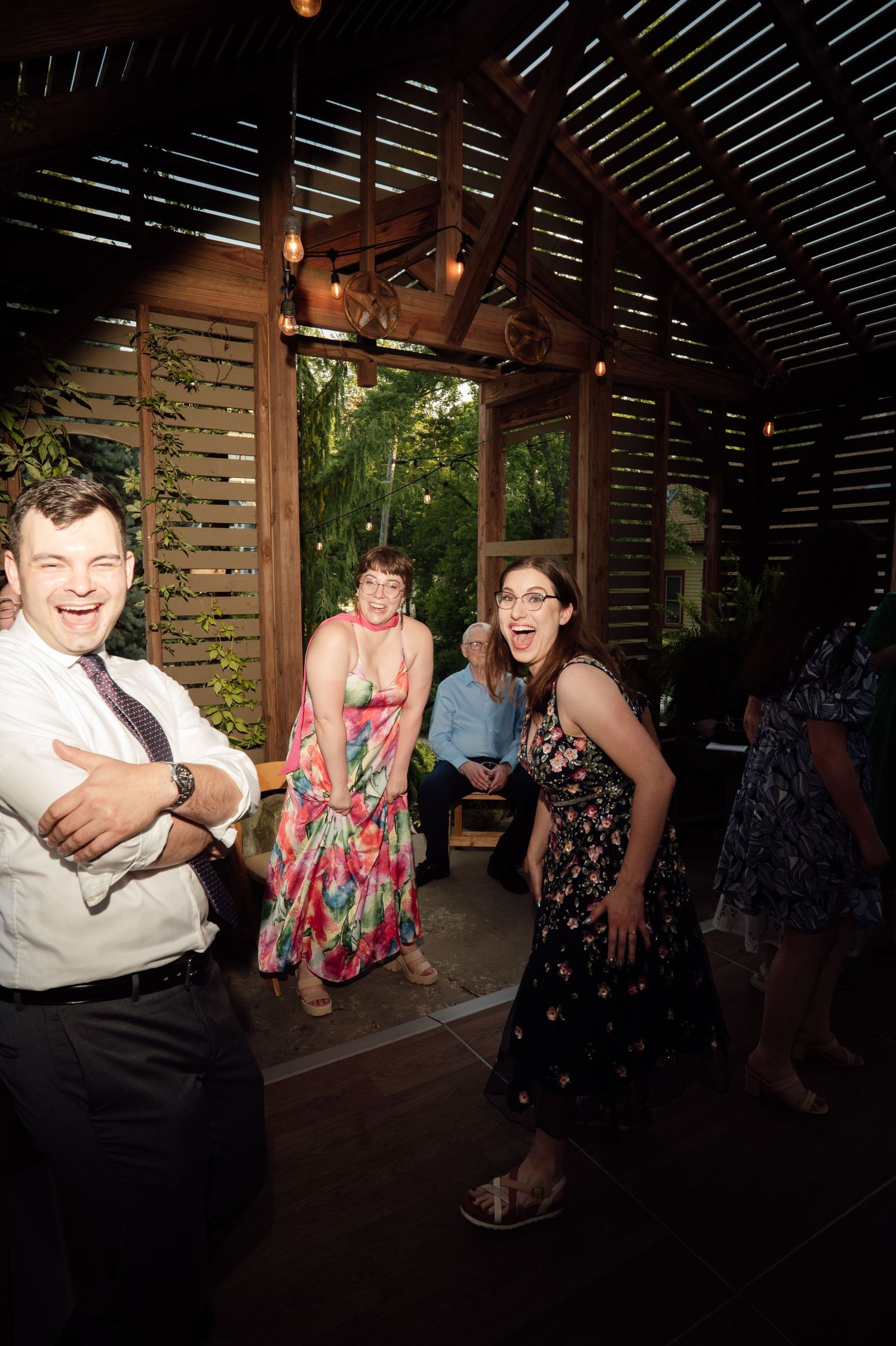 Guests laughing and dancing at an outdoor wedding reception in a rustic wooden pavilion with string lights.