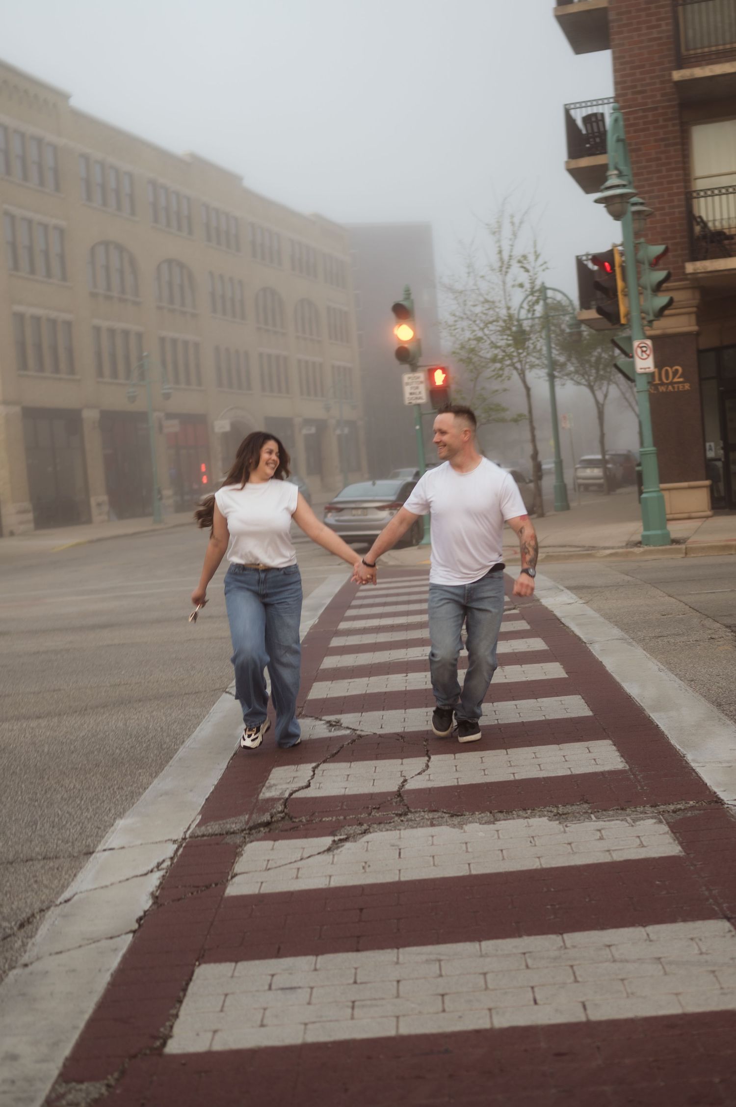 A couple holds hands while crossing a crosswalk on a misty morning in a downtown urban setting.